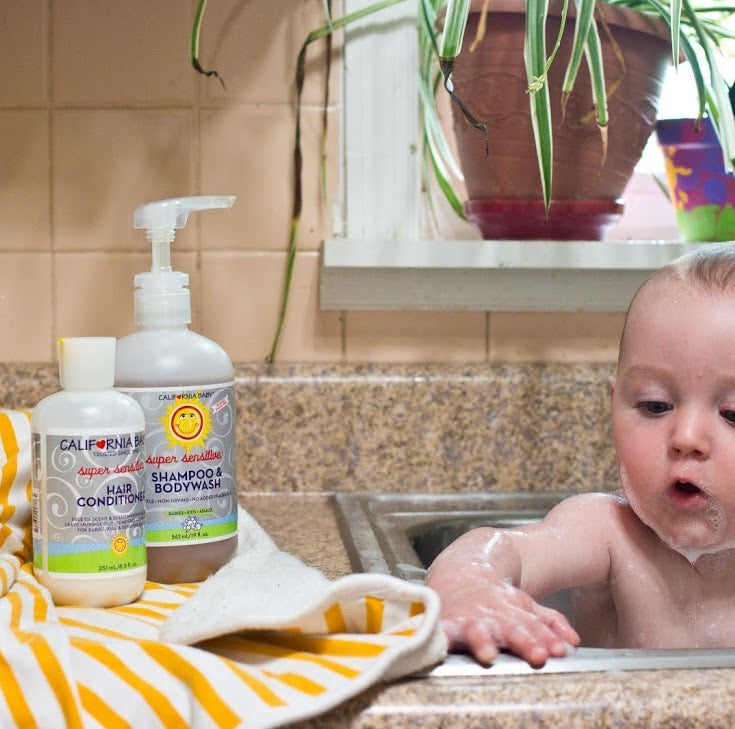 Baby in a bath with California Baby shampoo and bodywash bottles on a tiled bathroom counter. California Baby Super Sensitive Bath Essentials Bundle | Fragrance-Free Baby Shampoo, Body Wash, Conditioner, Calendula Cream, Lotion & Hand Sanitizer