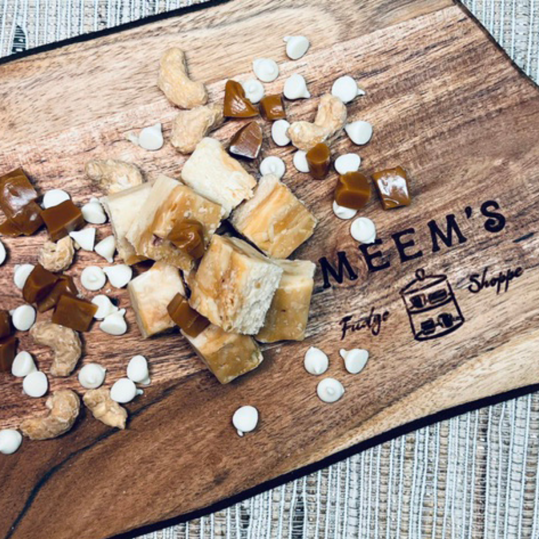 Handmade salted caramel cashew fudge cubes arranged on a rustic wooden cutting board engraved with Meem's Fudge Shoppe, surrounded by whole cashews, soft caramel pieces, and white chocolate chips on a woven placemat background.