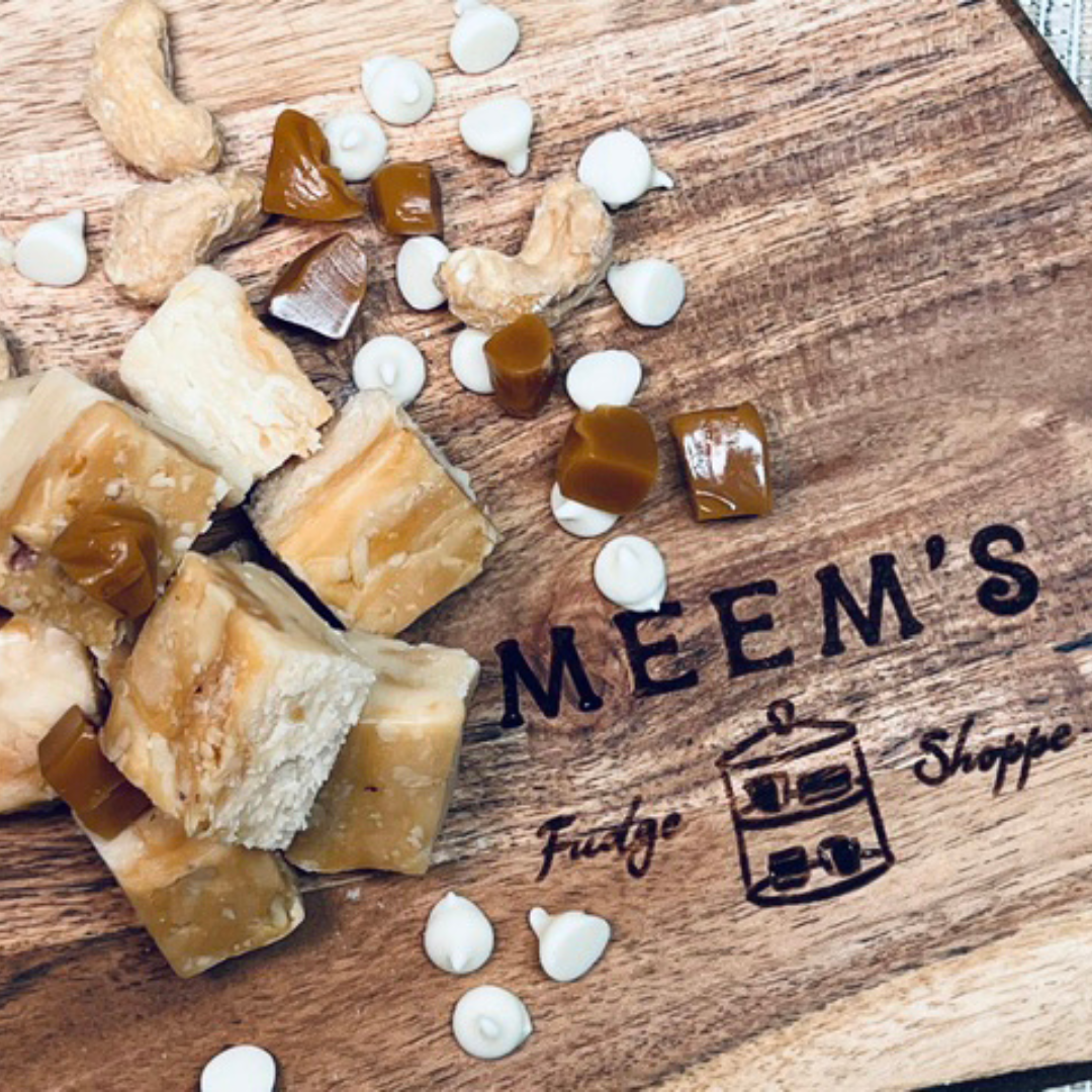 Handmade salted caramel cashew fudge cubes arranged on a branded wooden cutting board with scattered caramel pieces, whole cashews, and white chocolate chips.