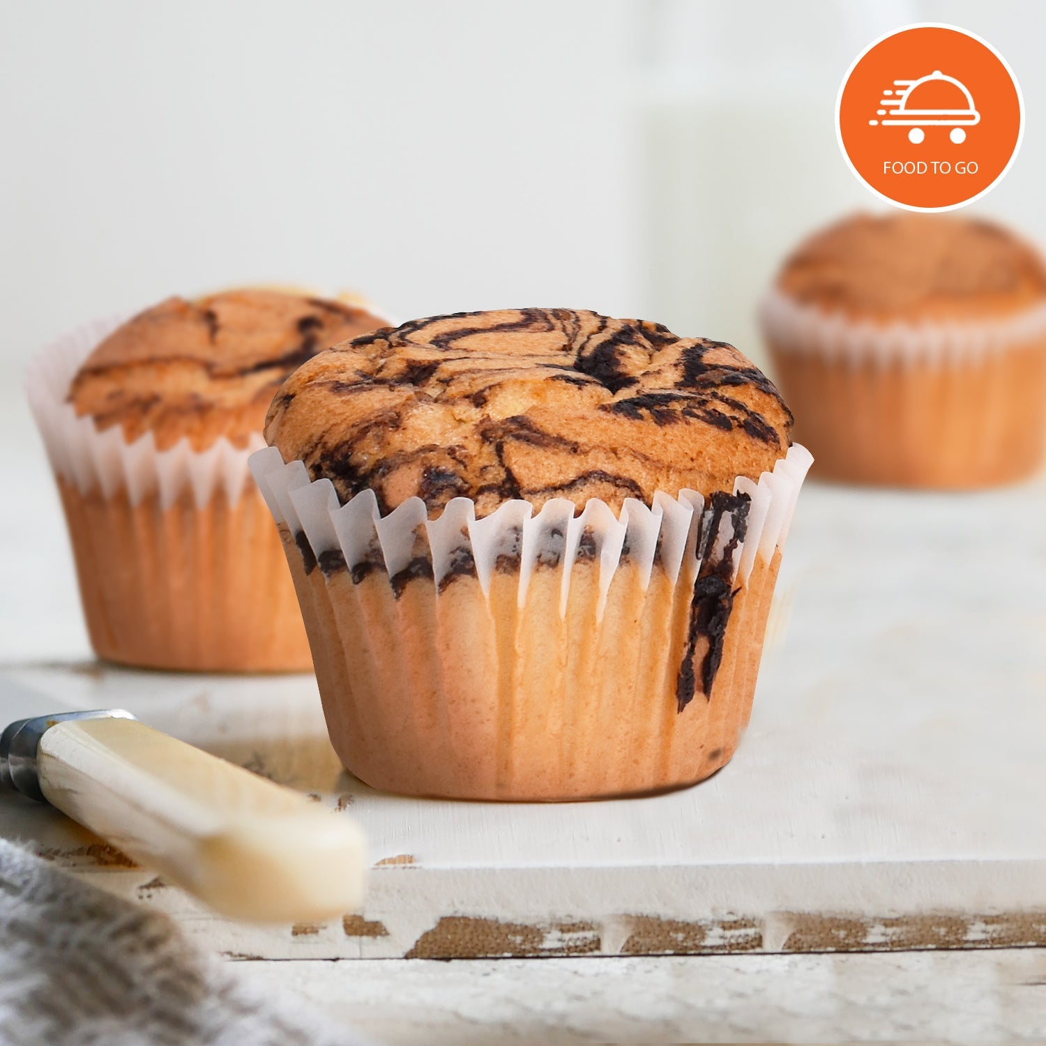 Marble swirl low-carb muffin in a white paper liner on a rustic wooden board, with two softly blurred muffins in the background and a butter knife in the foreground. Features a "Food To Go" logo badge in the upper-right corner. Ideal for keto, sugar-free, and low-carb breakfast or snack products.