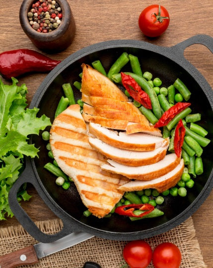 Grilled slices of certified organic air-chilled boneless skin-on chicken breast served over fresh green beans, peas, and red chili peppers in a black cast-iron skillet on a wooden table, garnished with lettuce, tomatoes, and a peppercorn grinder.