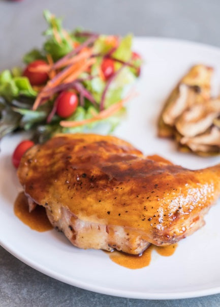 A roasted certified organic air-chilled boneless skin-on chicken breast with a golden-brown glazed surface, served on a white plate with mixed salad, sautéed mushrooms, and savory sauce on a gray tabletop.