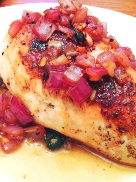 A close-up of a cooked certified organic air-chilled boneless skin-on chicken breast topped with diced red onions, garlic, and herbs in a savory sauce, served on a white plate.