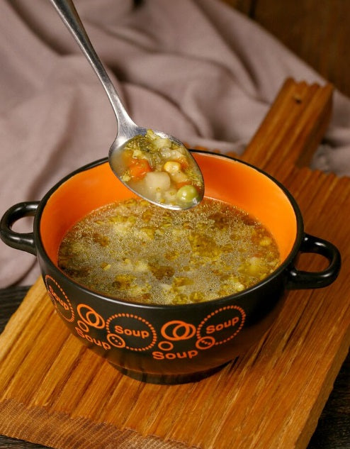 A black ceramic soup bowl with an orange interior, filled with a vegetable soup, resting on a wooden board. A silver spoon is shown lifting some of the soup, which contains green beans, carrots, and peas. Certified Organic Chicken Broth Bones | Non-GMO, Soy-Free, Non-Medicated | Hendricks Farm & Hatchery
