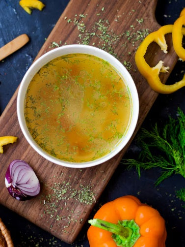 Golden bowl of homemade chicken bone broth garnished with herbs, served on a rustic wooden board with fresh vegetables. Certified organic chicken broth bones from Hendricks Farm & Hatchery, non-GMO, soy-free, and non-medicated for making rich, nutritious homemade stock.