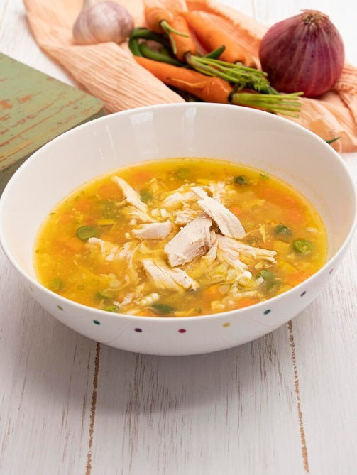 A white ceramic bowl filled with chicken soup and vegetables, set on a white wooden table with carrots, garlic, and an onion blurred in the background. Certified Organic Chicken Broth Bones | Non-GMO, Soy-Free, Non-Medicated | Hendricks Farm & Hatchery