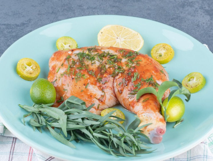 A cooked organic chicken leg quarter garnished with fresh herbs and citrus slices, served on a light blue oval plate against a gray background. Certified Organic Pasture-Raised, Non-GMO, Soy-Free.