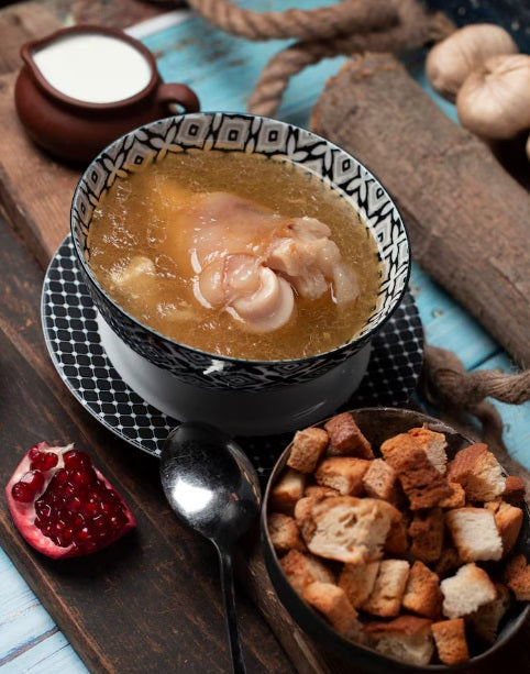 A bowl of chicken soup with a black and white patterned rim, served on a matching saucer with a spoon, alongside a bowl of croutons, a pomegranate, and rustic wooden props on a blue table. Certified Organic Chicken Broth Bones | Non-GMO, Soy-Free, Non-Medicated | Hendricks Farm & Hatchery