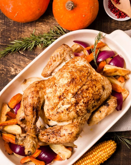 A whole roasted USDA Organic pasture-raised chicken in a white oval baking dish with roasted carrots, red onions, and mushrooms, garnished with fresh rosemary and surrounded by pumpkins and corn on a rustic wooden table.