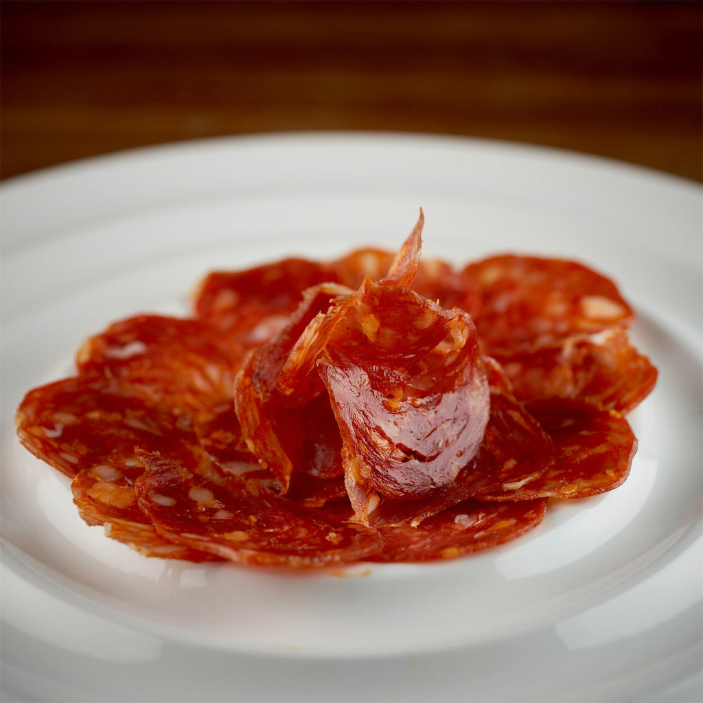 Thin slices of premium Ibérico chorizo artfully arranged on a white plate, showcasing rich marbling and deep red color against a softly blurred wooden background.