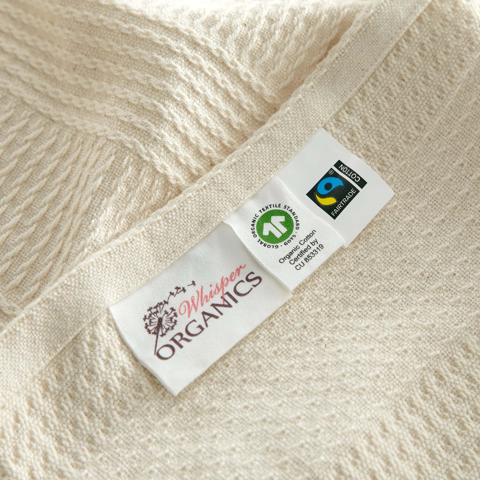 Close-up of a label on a beige fabric with 'Whisper Organics' branding. Organic Cotton Throw with Tassels