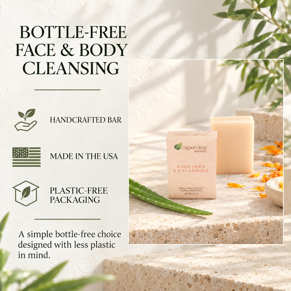 A bottle-free face and body cleansing bar set by Open Kay Organics, featuring a small rectangular bar of soap in a paper sleeve and a larger matching bar behind it, styled on a stone surface with aloe vera and orange petals. Text on the left highlights handcrafted bar, made in the USA, and plastic-free packaging.