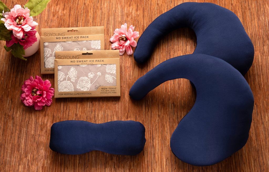 Navy blue U-shaped neck support pillow and rectangular ice packs on a rustic wooden surface with pink flowers, featuring a soothing self-care comfort setup for post-surgery recovery, mastectomy care, breast cancer recovery, pain relief, cold therapy, neck support, wellness, comfort, and thoughtful recovery gifting.