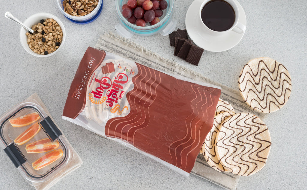 Overhead view of Kim's Magic Pop dark chocolate drizzled rice cakes in opened packaging with coffee, grapes, granola, walnuts, orange slices, and dark chocolate pieces on a light countertop.