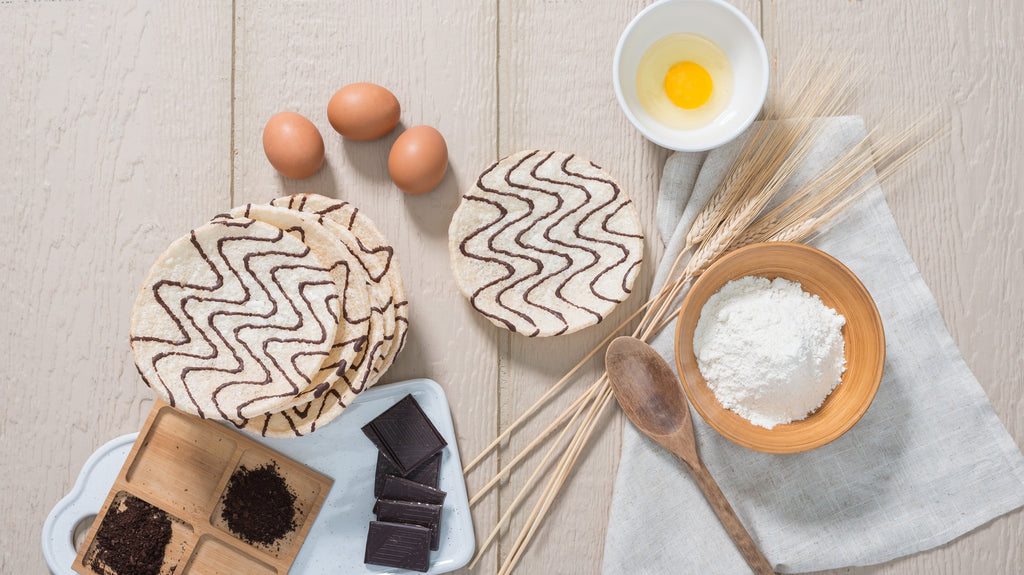 Baking ingredients including flour, eggs, wheat stalks, dark chocolate, and wooden spoon arranged beside Kim's Magic Pop drizzled rice cakes on a light wooden surface.