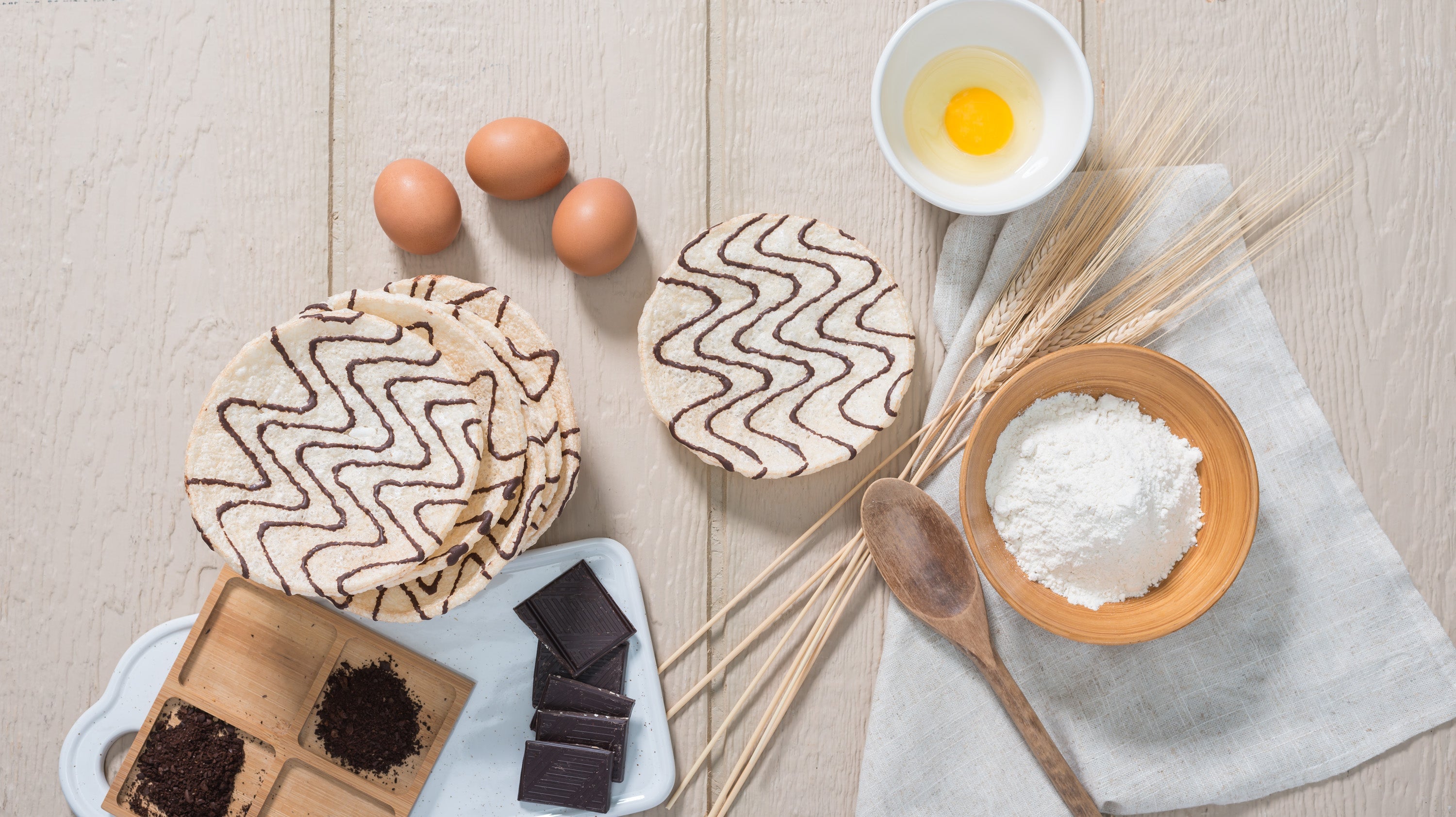 Baking ingredients including flour, eggs, wheat stalks, dark chocolate, and wooden spoon arranged beside Kim's Magic Pop drizzled rice cakes on a light wooden surface.