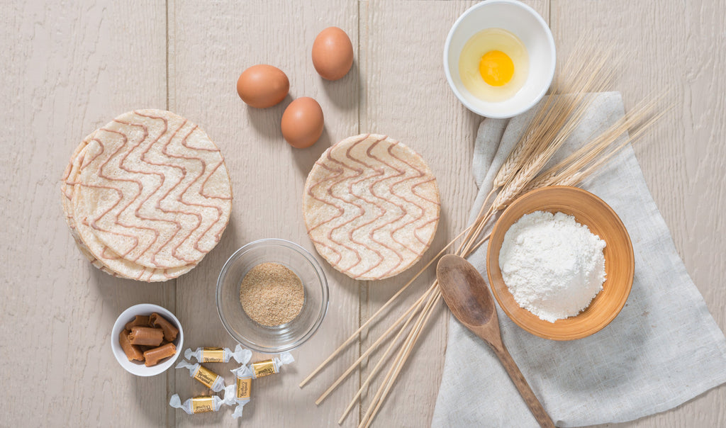 Overhead view of Kim's Magic Pop drizzled rice cakes surrounded by baking ingredients including eggs, flour, wheat stalks, raw sugar, caramel candies, and a wooden spoon on a light wooden surface.