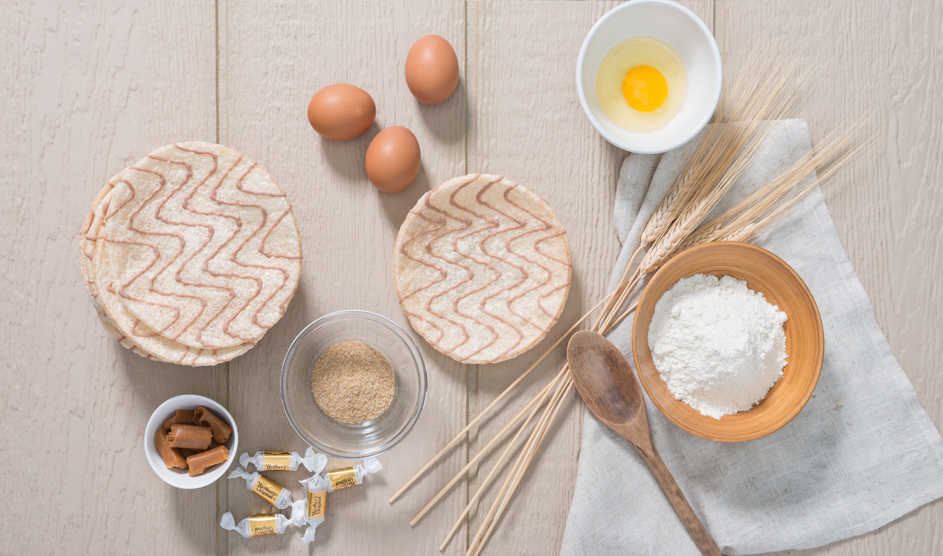 Overhead view of Kim's Magic Pop drizzled rice cakes surrounded by baking ingredients including eggs, flour, wheat stalks, raw sugar, caramel candies, and a wooden spoon on a light wooden surface.