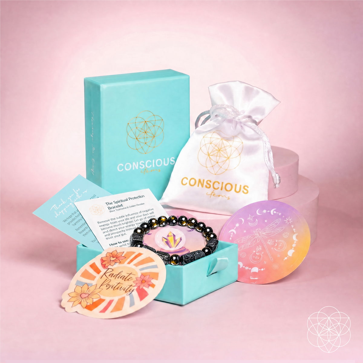 A Conscious Living self-care gift set arranged on a pink studio background. The set includes a turquoise branded box, a white satin drawstring pouch, a black beaded bracelet, a pastel gradient soap bar, a card, and a decorative tag. Direction of Wealth & Success - The Spiritual Protection Bracelet with Black Tourmaline and Golden Obsidian for Absorbing Negativity
