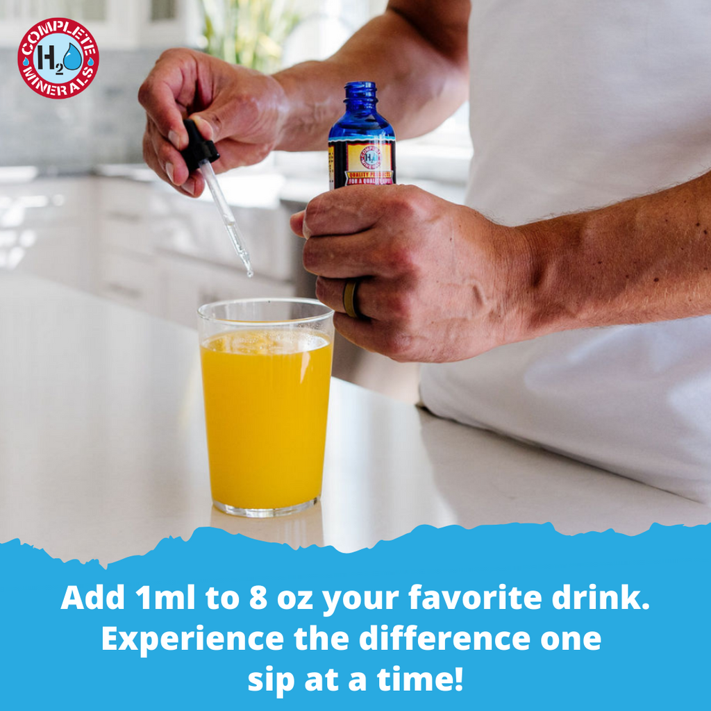 Person adding a drop of a blue bottle into a glass of orange juice with a white background.