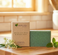 A bar of Aspen Kay Naturals Dead Sea Mud & Neem Leaf soap stands in front of its kraft paper packaging box on a wooden surface, styled with mint leaves and small white flowers against a warm, blurred bathroom background.