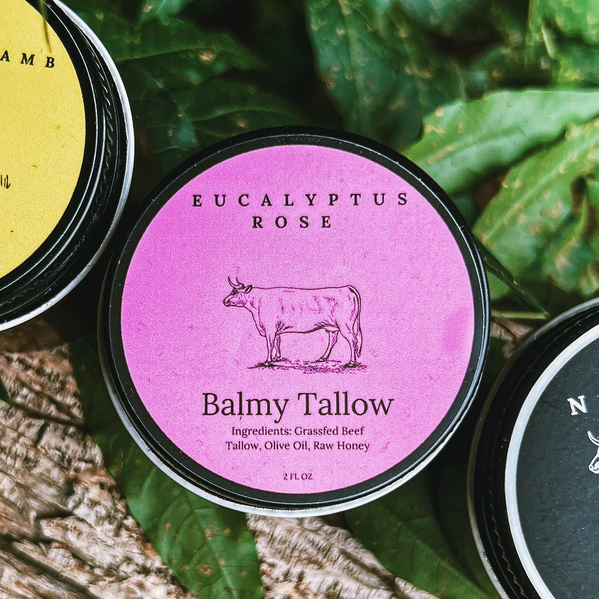 A round tin of Balmy Tallow Eucalyptus Rose body butter is centered on a bed of green leaves and wood, with other tins partially visible at the left and right edges. Eucalyptus Rose Tallow Balm 4 oz. | Nourishing Floral Skincare | Balmy Tallow