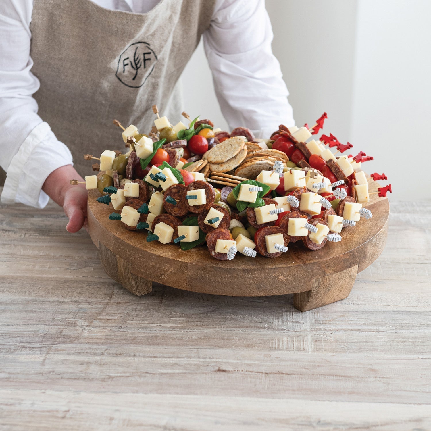 Person holding a wooden platter with a colorful assortment of snacks on a light wooden surface. French Saucisson Sec Salumi | Finch + Fennel