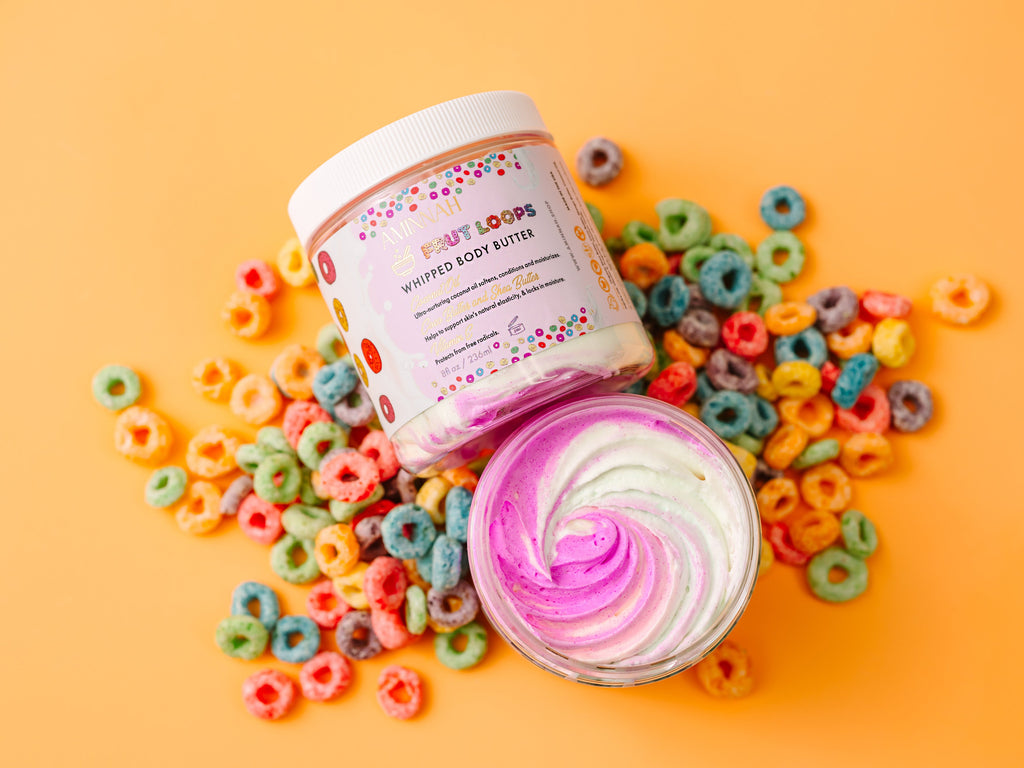 Body butter with colorful packaging surrounded by cereal on a yellow background. AMINNAH Frut Loops Whipped Body Butter opened to reveal a vibrant pink and white whipped texture, displayed on a bright yellow background surrounded by colorful cereal pieces. Clean beauty, cruelty-free, vegan body butter made with naturally derived ingredients including shea butter, coconut oil, jojoba oil, avocado oil, sweet almond oil, and cocoa butter to deeply nourish, soften, and hydrate skin. Female-owned brand available