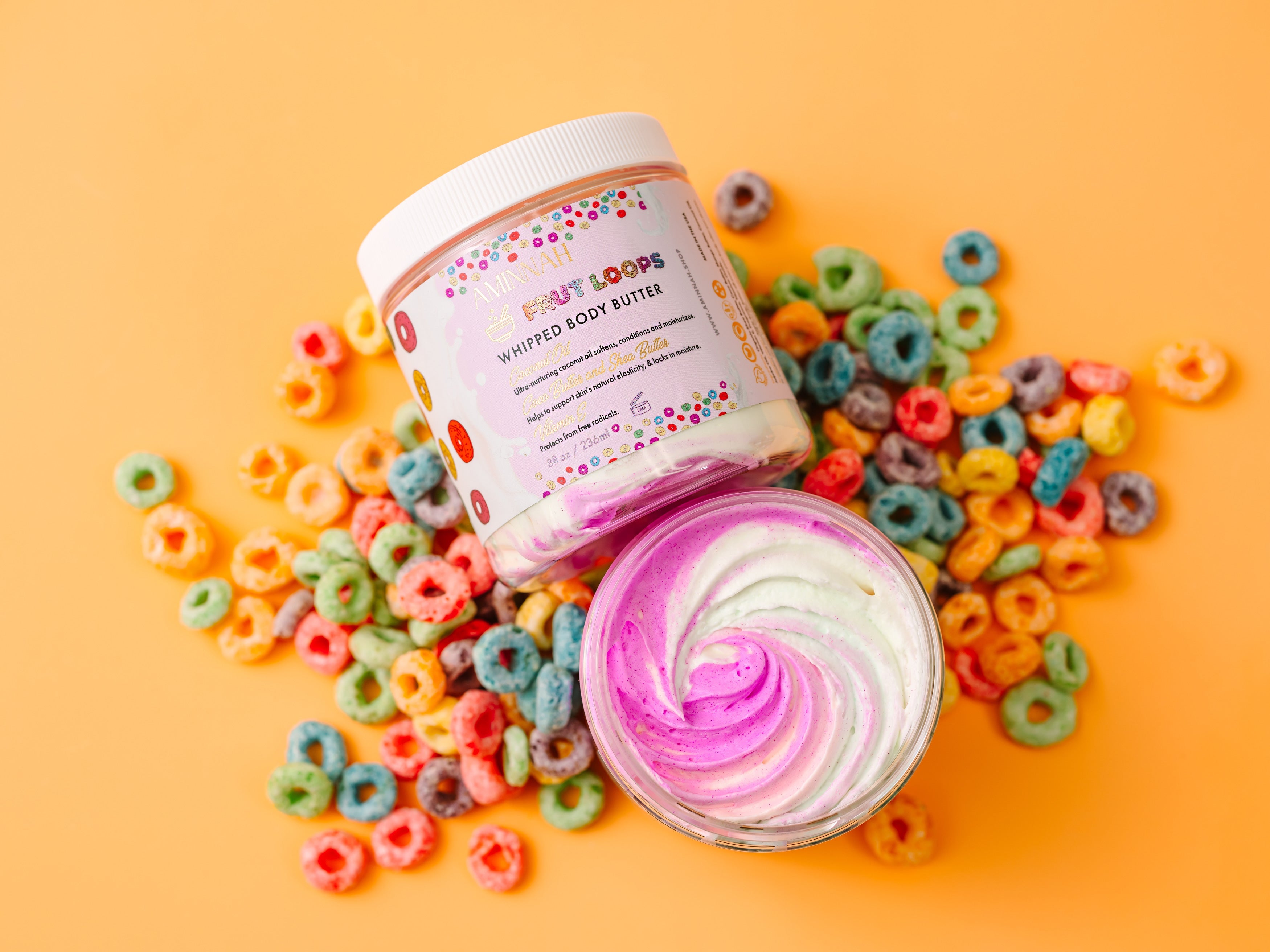 Body butter with colorful packaging surrounded by cereal on a yellow background. AMINNAH Frut Loops Whipped Body Butter opened to reveal a vibrant pink and white whipped texture, displayed on a bright yellow background surrounded by colorful cereal pieces. Clean beauty, cruelty-free, vegan body butter made with naturally derived ingredients including shea butter, coconut oil, jojoba oil, avocado oil, sweet almond oil, and cocoa butter to deeply nourish, soften, and hydrate skin. Female-owned brand available