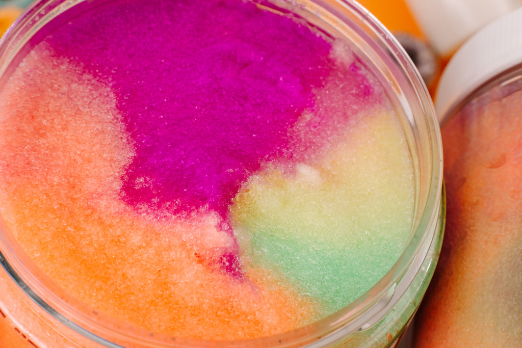 Colorful body scrub in a jar with a bottle of liquid soap in the background. Close-up of AMINNAH Frut Loops Sugar Polish showcasing its vibrant pink, orange, yellow, and green exfoliating sugar crystals. Clean beauty, cruelty-free, vegan sugar scrub made with naturally derived ingredients to gently exfoliate, remove dull skin, and reveal a softer, smoother, more radiant complexion. Female-owned brand available at Tree of Life Grocers.