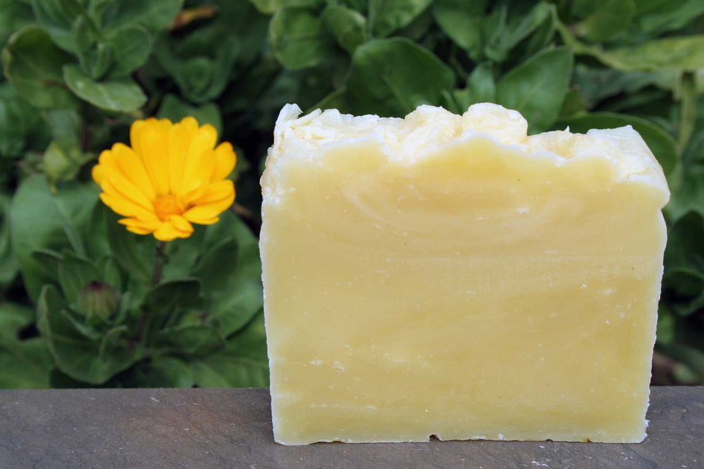Bar of yellow soap with a white lather on top, placed on a surface with green foliage and a yellow flower in the background.