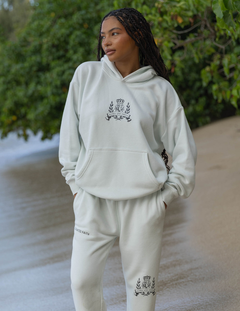 Person wearing a light gray hoodie and pants with an emblem on a beach.