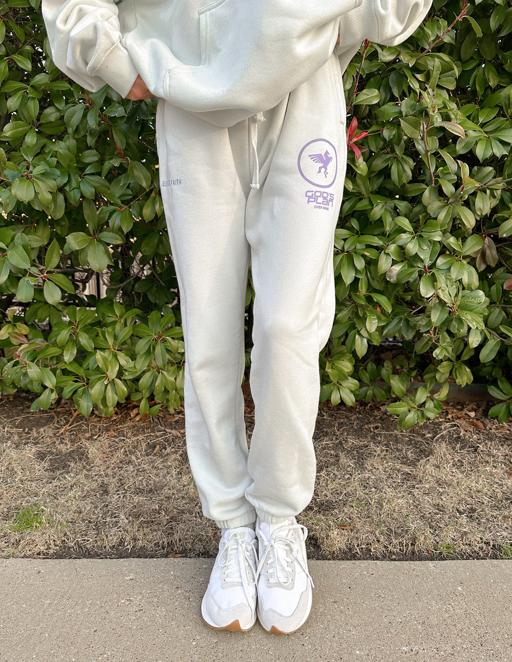 Person wearing light gray sweatpants with a logo, standing outdoors against green bushes.