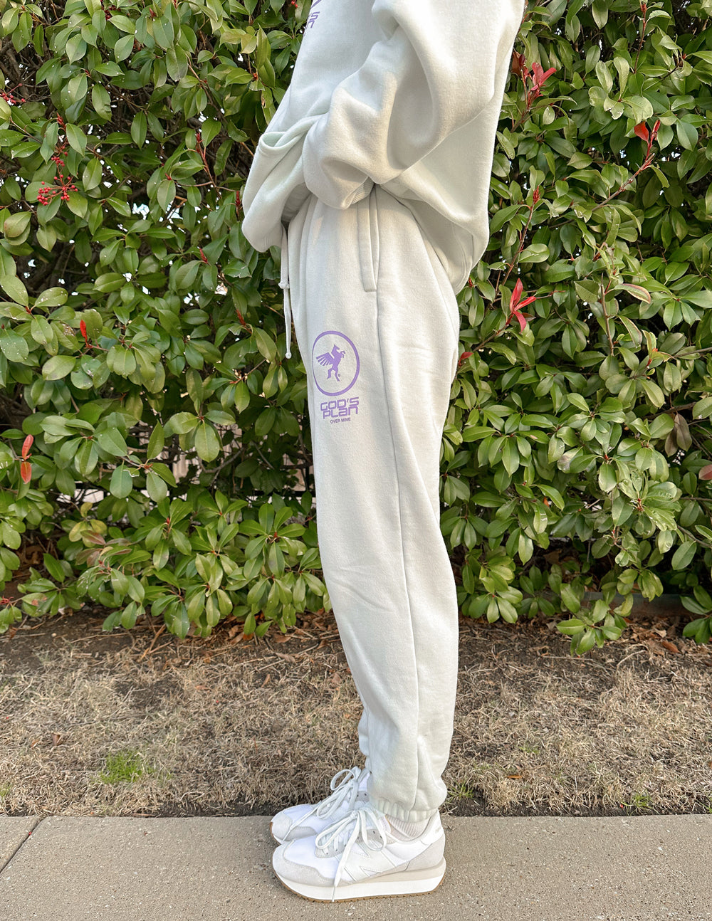 Person wearing a light gray tracksuit with a logo in front of green bushes