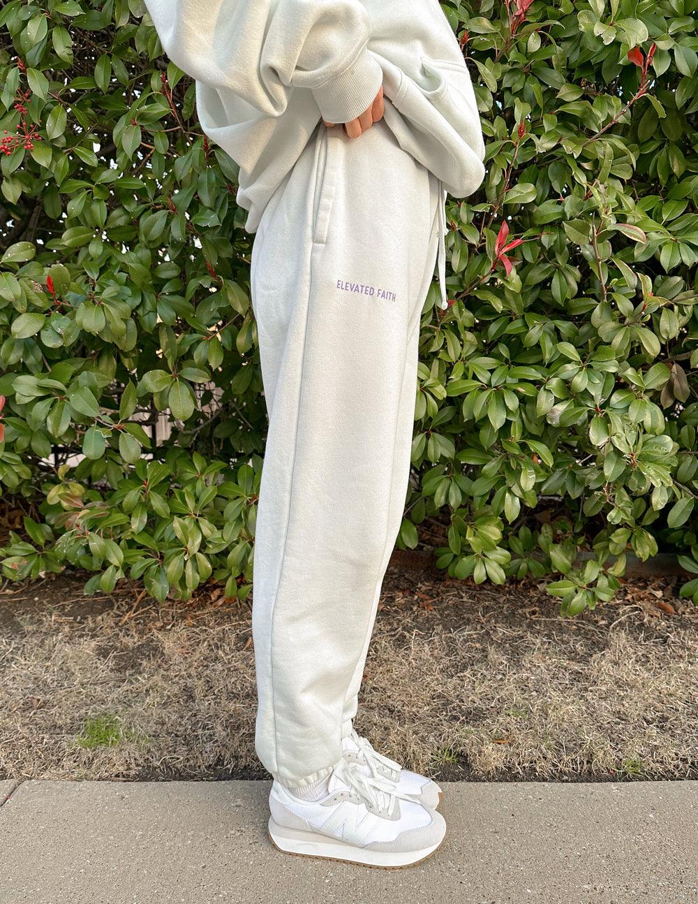 Person wearing light gray sweatpants with a brand logo, standing against a green bush.