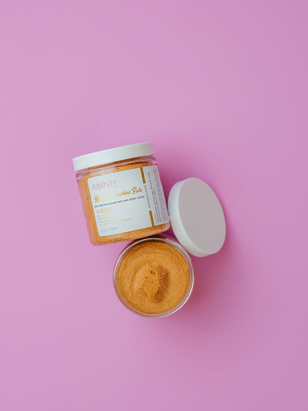 Golden Sunshine Babe Sugar Scrub by AMINNAH on pink background with open jar showcasing golden exfoliating sugar scrub. Clean beauty, cruelty-free, vegan, paraben-free, gluten-free body scrub made with natural ingredients for smooth, radiant skin. Female-owned brand with recyclable packaging, luxury skincare and self-care body polish.