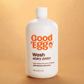 A 16 fl oz bottle of Good Egg Wash Heavy Doody plant-based enzyme cleanser, featuring a white speckled bottle with orange and brown text, set against a warm tan background.