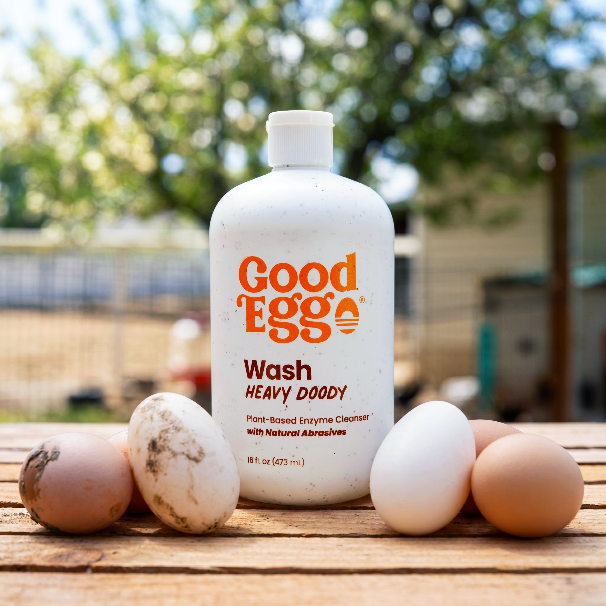 A white bottle of Good Egg Wash Heavy Doody egg cleaner stands on a wooden table outdoors, surrounded by several eggs of different colors. The background is a soft-focus farm setting with green foliage.