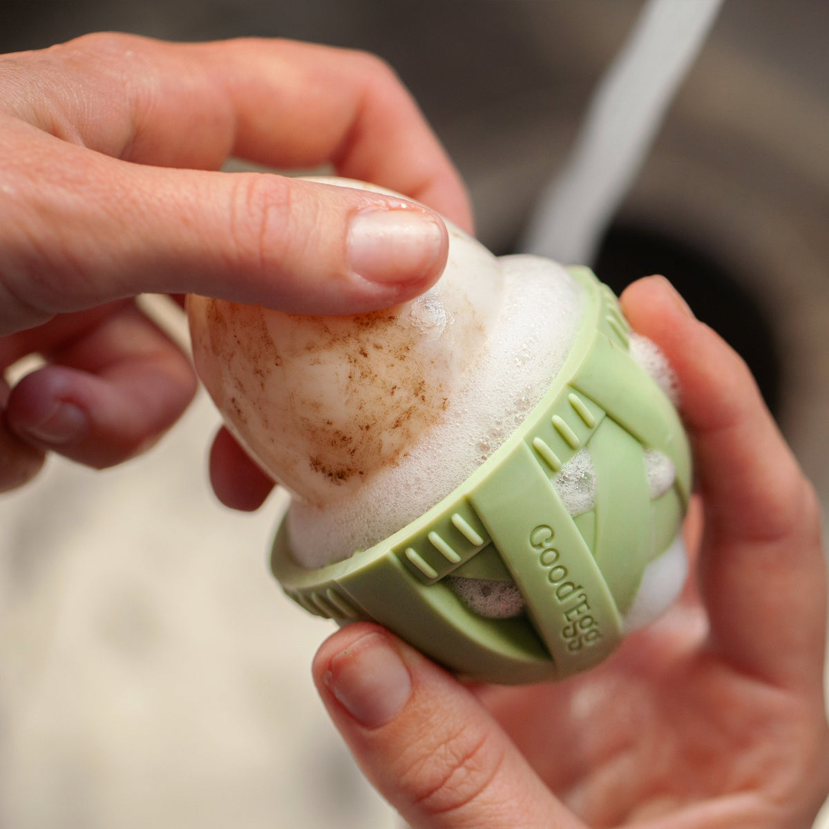 Fresh egg being cleaned with a foaming green GoodEgg silicone egg scrubber, showing gentle removal of dirt from the shell under running water.