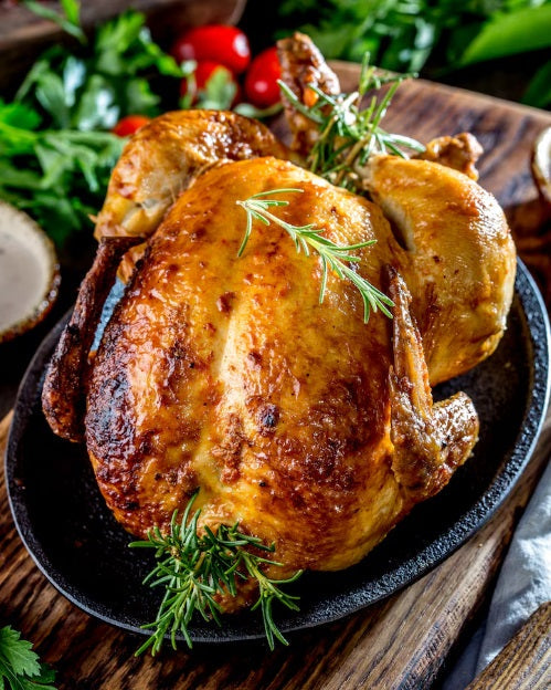 A whole roasted chicken with crispy golden-brown skin, garnished with fresh rosemary sprigs, served on a black cast-iron skillet over a rustic wooden table with fresh herbs and tomatoes in the background.