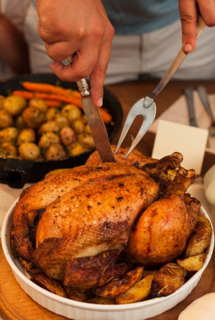 A roasted USDA Organic whole chicken in a white oval baking dish with roasted potatoes, being carved with a knife and carving fork. Pasture-raised, Non-GMO, soy-free chicken from Hendricks Farm & Hatchery, vacuum sealed for freshness.