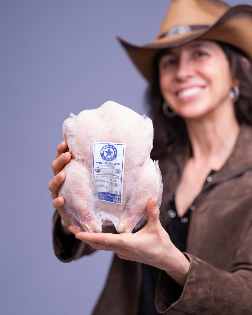 Person holding a vacuum-sealed USDA Organic whole chicken with the Hendricks Farm & Hatchery label, wearing a cowboy hat against a gray background. Pasture-raised, non-GMO, soy-free whole chicken package.