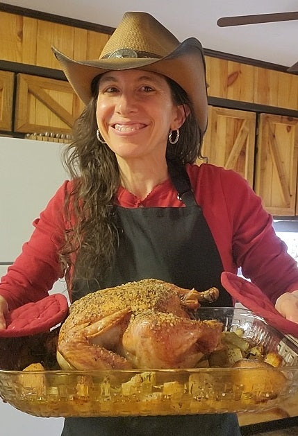 A smiling woman wearing a cowboy hat, red shirt, and black apron proudly holds a glass baking dish with a golden roasted whole chicken in a rustic kitchen with wooden cabinets, showcasing a home-cooked farm-style meal.