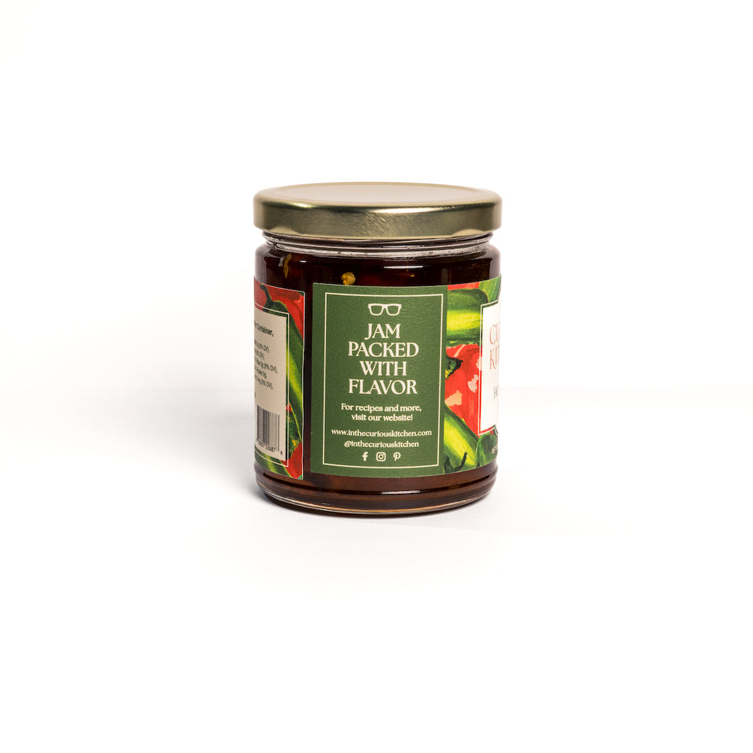 Back view of In the Curious Kitchen Hot Pepper Jelly jar showing the product label with “Jam Packed With Flavor,” gold lid, and sweet and spicy pepper jelly in a small glass jar on a white background.