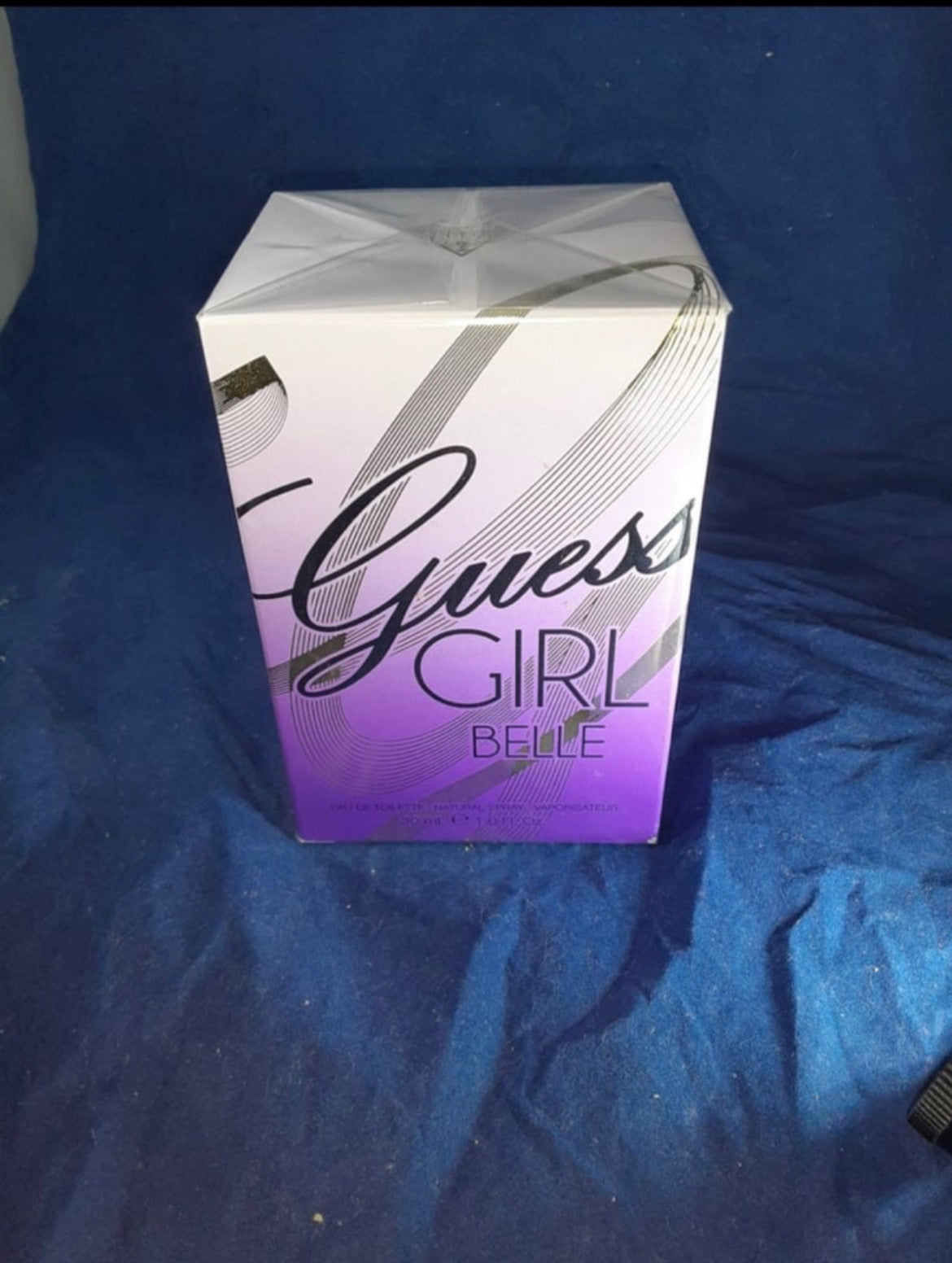 Guess Girl Belle perfume box with a purple and white gradient design, placed on a blue fabric background.