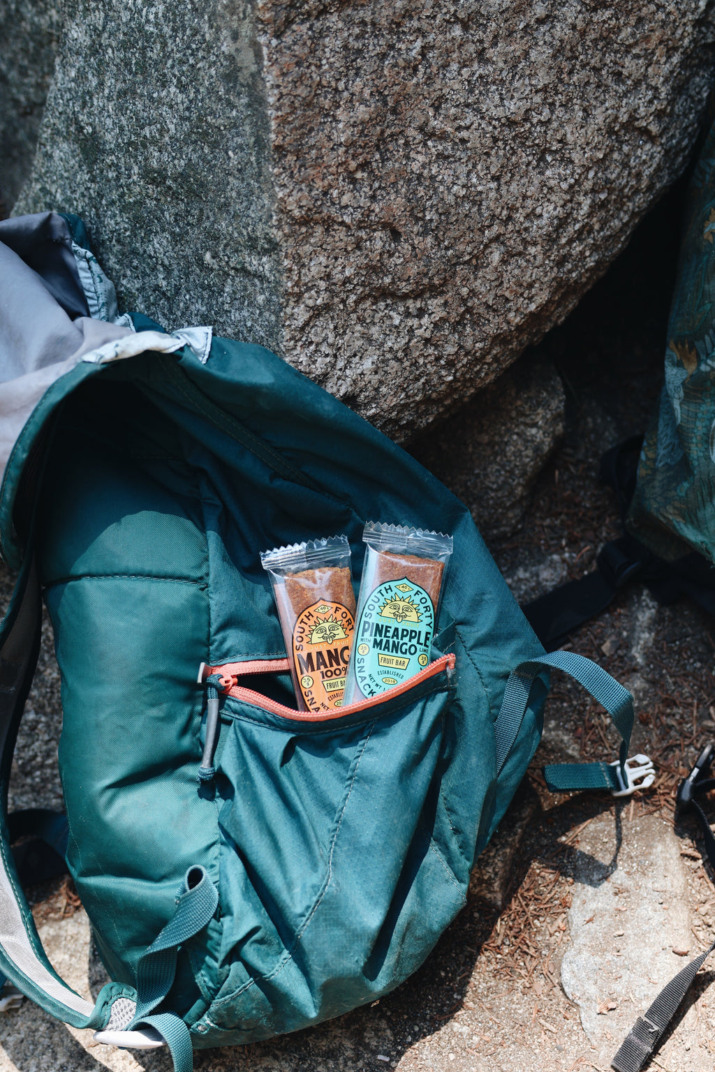 South Forty 100% Mango fruit bars tucked into the front pocket of a teal hiking backpack on a rocky mountain trail, healthy vegan gluten-free one-ingredient dried mango snacks, portable outdoor adventure snack, tropical fruit bars, natural non-GMO fruit snacks, hiking backpack essentials, Product of Ghana and Ivory Coast.