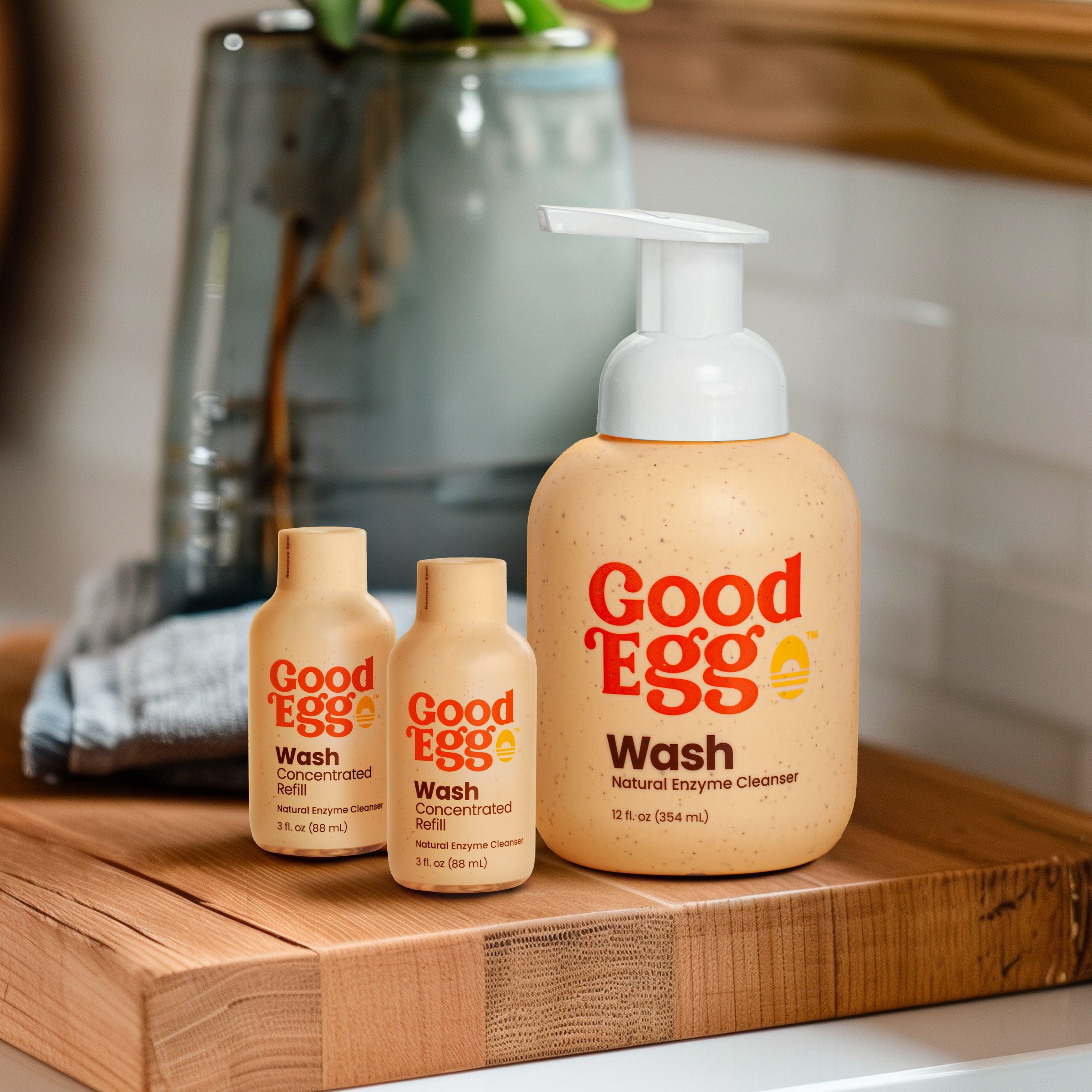 GoodEgg Wash natural enzyme egg cleanser bottles, including a 12 oz foaming pump bottle and two concentrated refill bottles, displayed on a wooden countertop in a farmhouse kitchen.