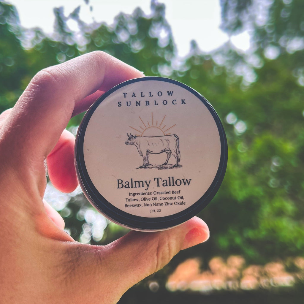 A hand holds a round black-lidded jar of Tallow Sunblock Balmy Tallow outdoors against a blurred green foliage background. The label features a cow illustration and text including 'TALLOW SUNBLOCK' and 'Balmy Tallow'. Ingredients: Grassfed Beef Tallow, Olive Oil, Coconut Oil, Beeswax, Non Nno Zinc Oxide