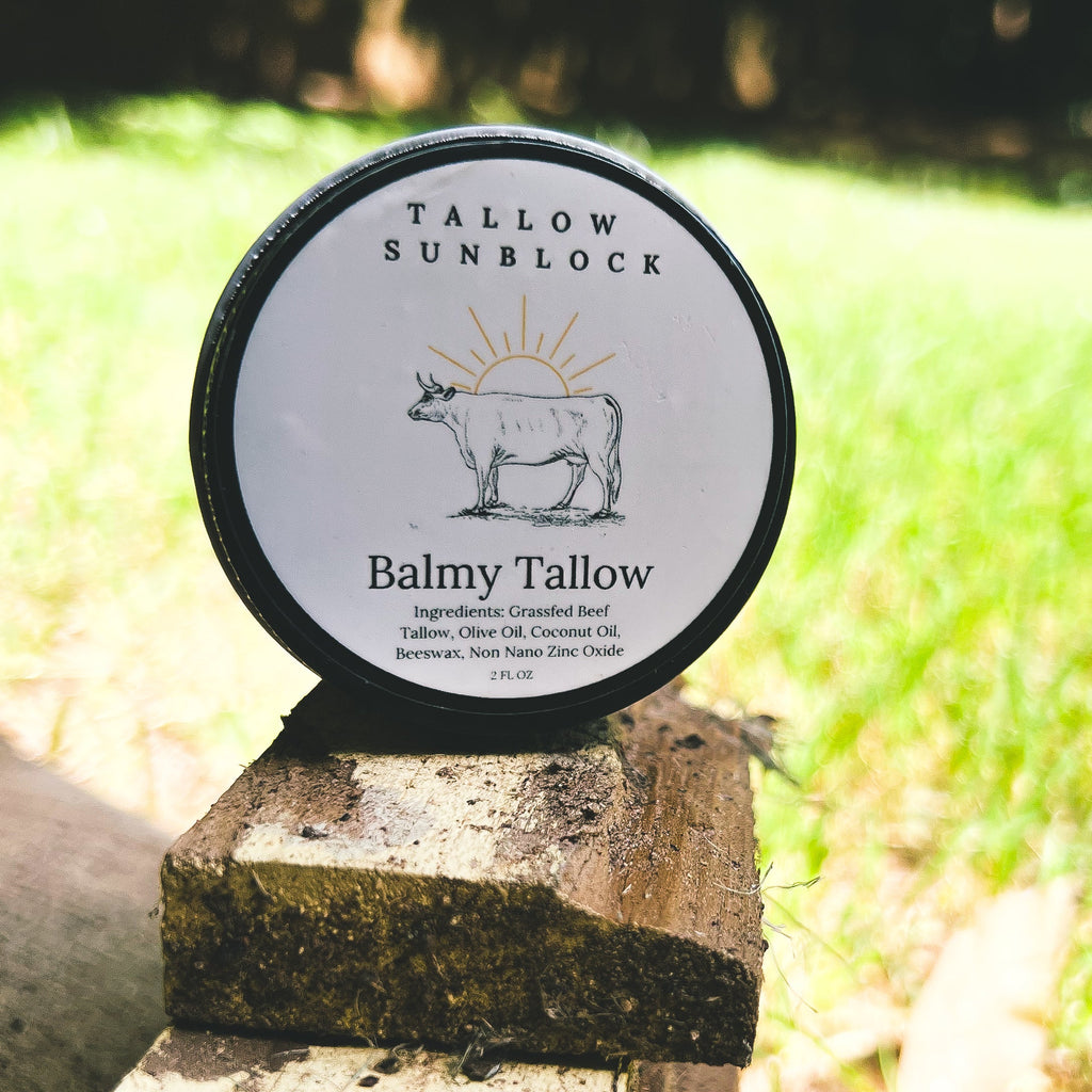 A round tin of Tallow Sunblock Balmy Tallow resting on a rustic wooden post outdoors, with a blurred green grassy background. Grassfed Beef Tallow, Olive Oil, Coconut Oil, Beeswax, Non Nno Zinc Oxide