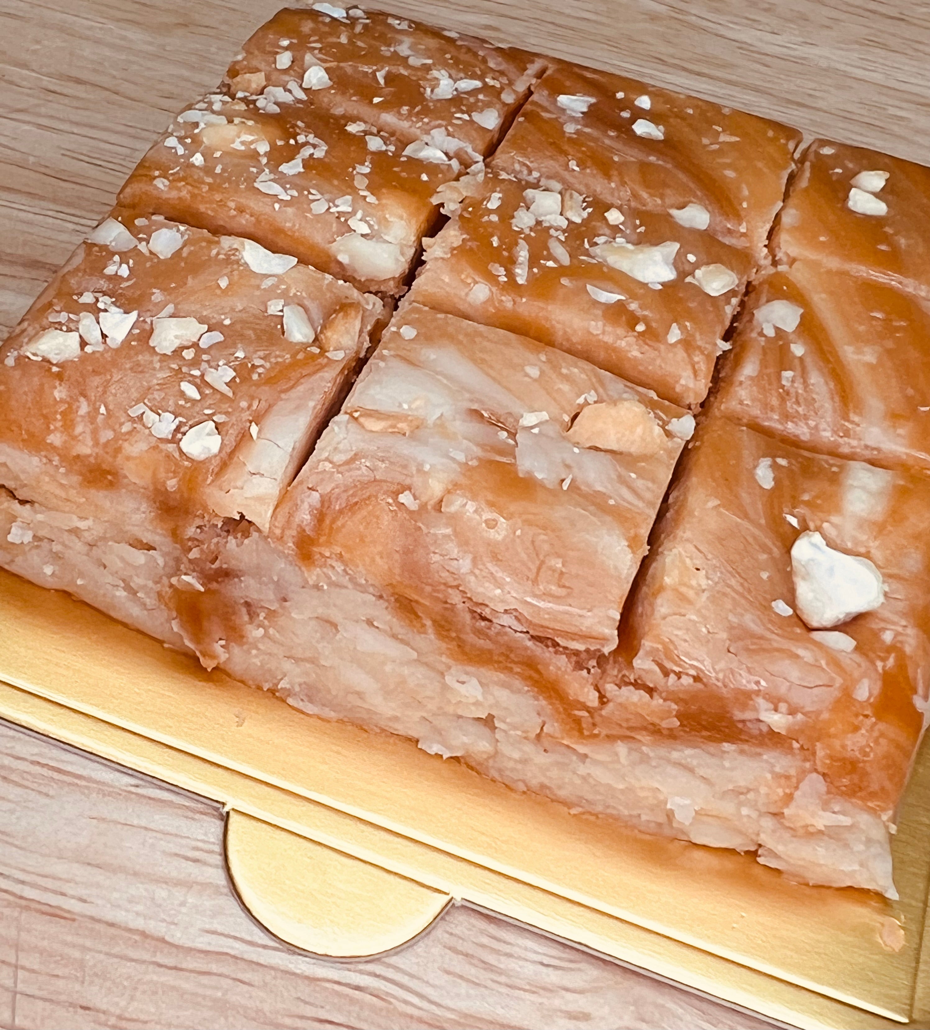 Handmade salted caramel cashew fudge cut into bite-sized squares and topped with chopped cashews, displayed on a gold serving tray over a light wood surface.